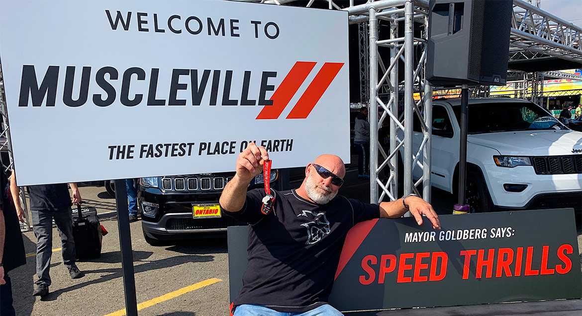 Bill Goldberg holding up his red key sitting on a bench that says "Mayor Goldberg Says: Speed Thrills" with a sign in the background that says "Welcome to Muscleville The Fastest Place on Earth"