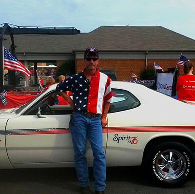 a man wearing an american flag patterned shirt standing in front of a spirit ‘76 edition Dodge Dart 