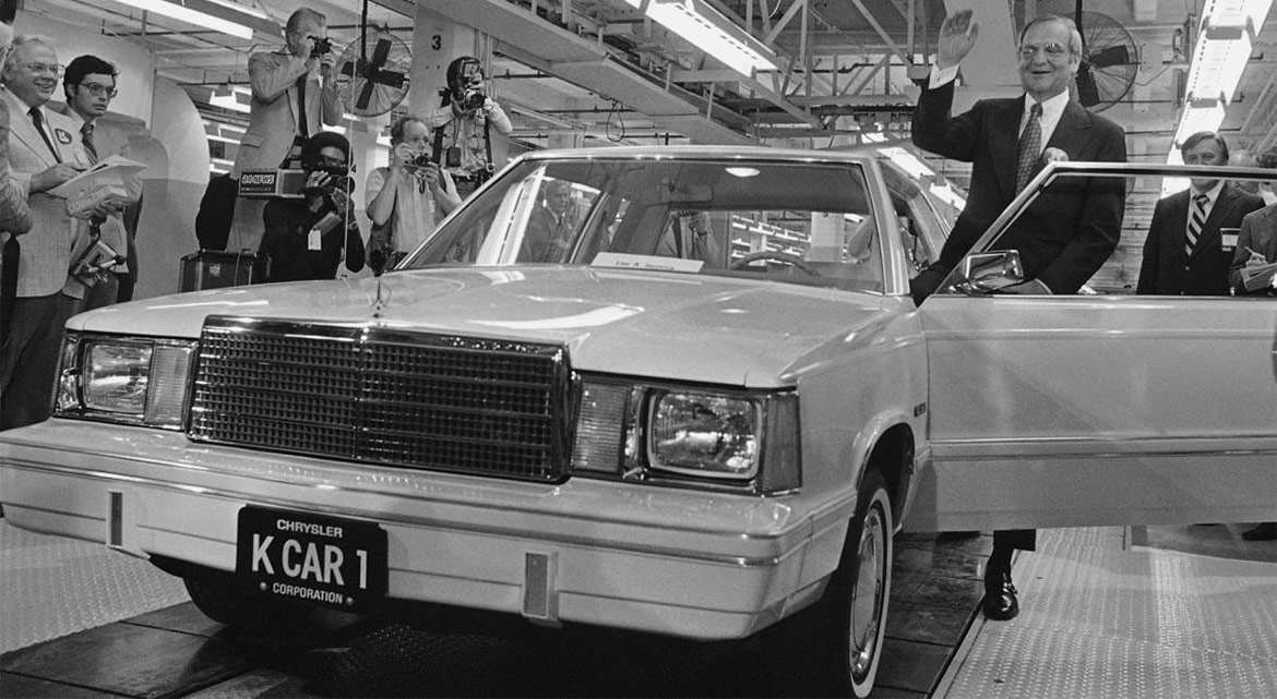 lee iococca standing in front of a car coming off the end of an assembly line