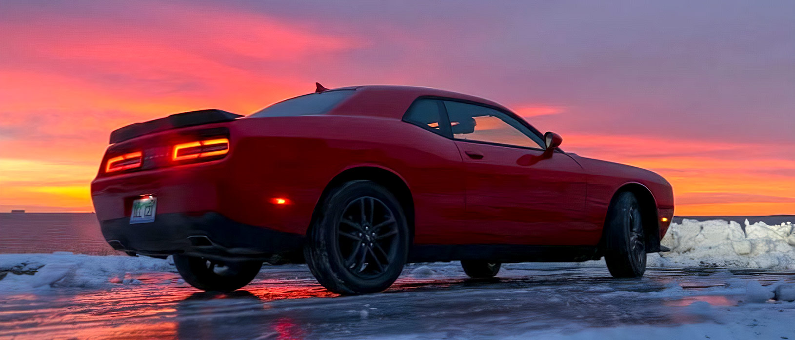 Red Dodge Challenger AWD parked in snow with purple and orange sunset behind