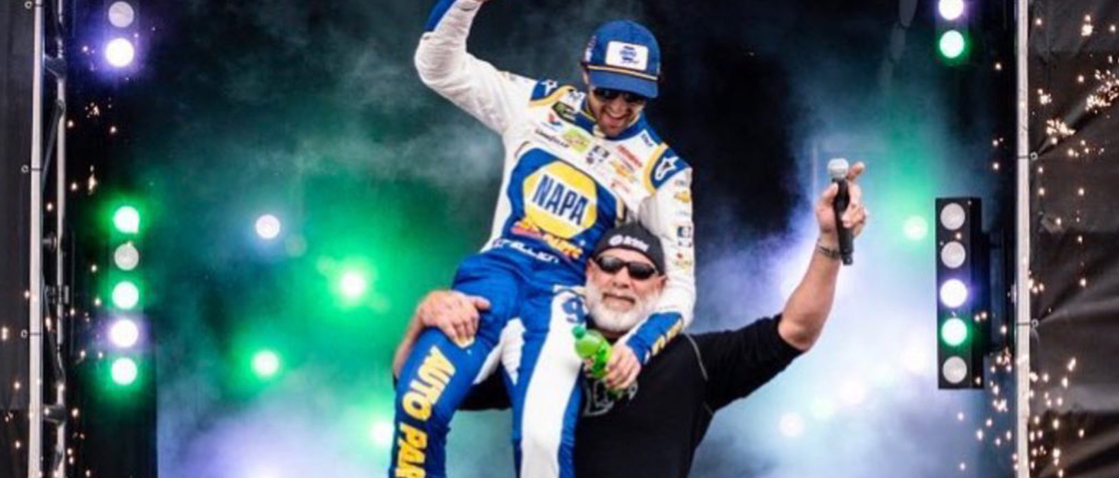 Goldberg holding up NASCAR driver with lights and fireworks in the background