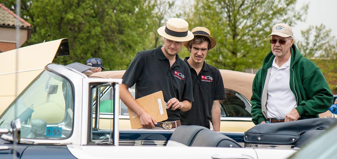 Two young men in hats speaking looking at a old car while a man in a green jacket stands next to them.