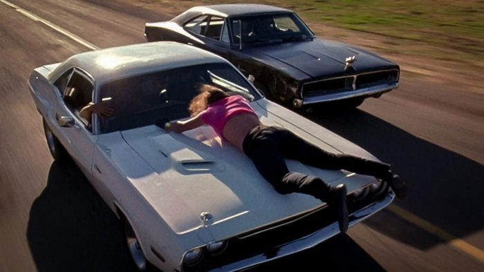 a man hanging onto the hood of a vehicle as it, and another vehicle, drive at high speeds