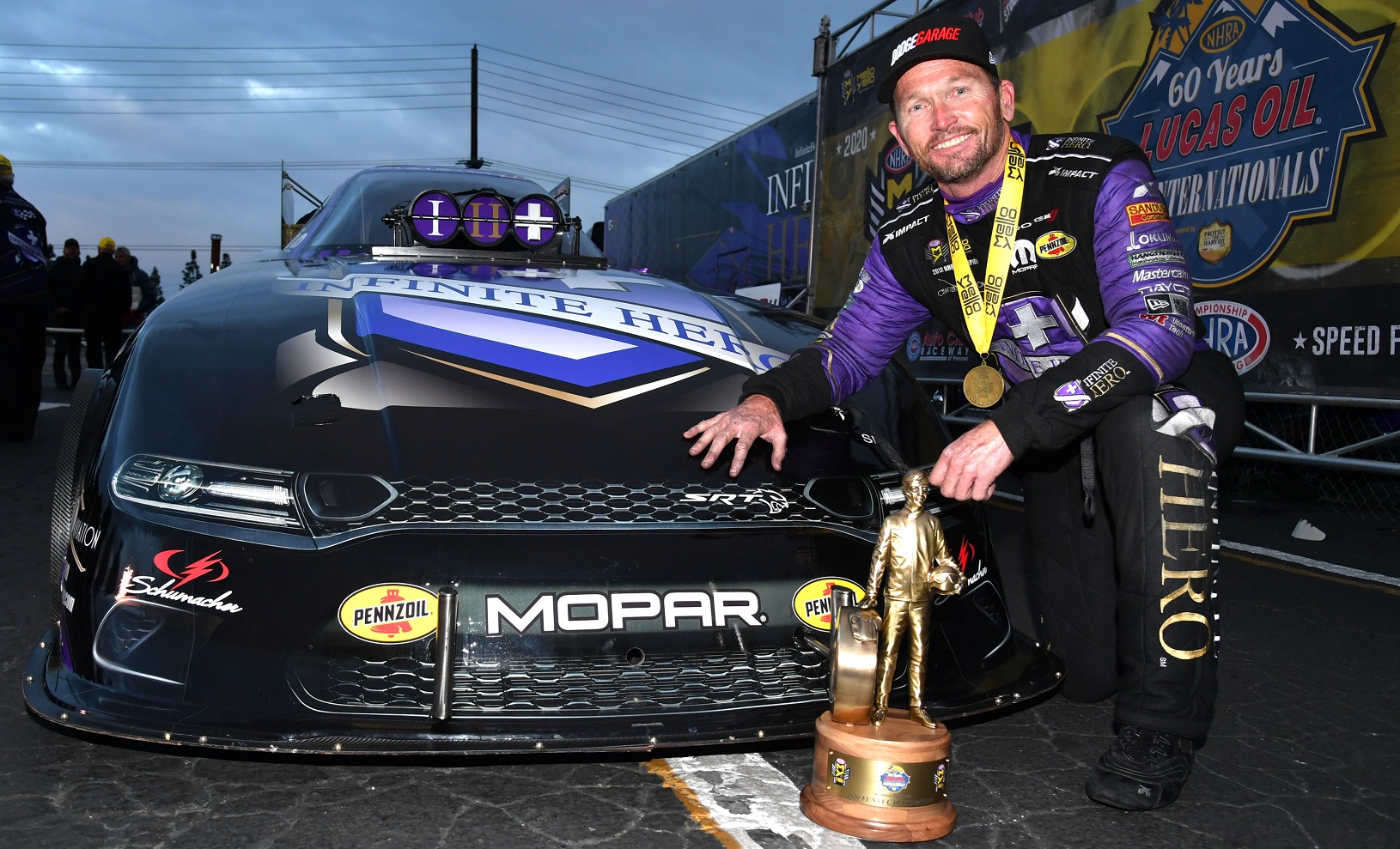 Jack Beckman kneeling in front of his Funny Car with the Wally trophy he won at NHRA Winternationals