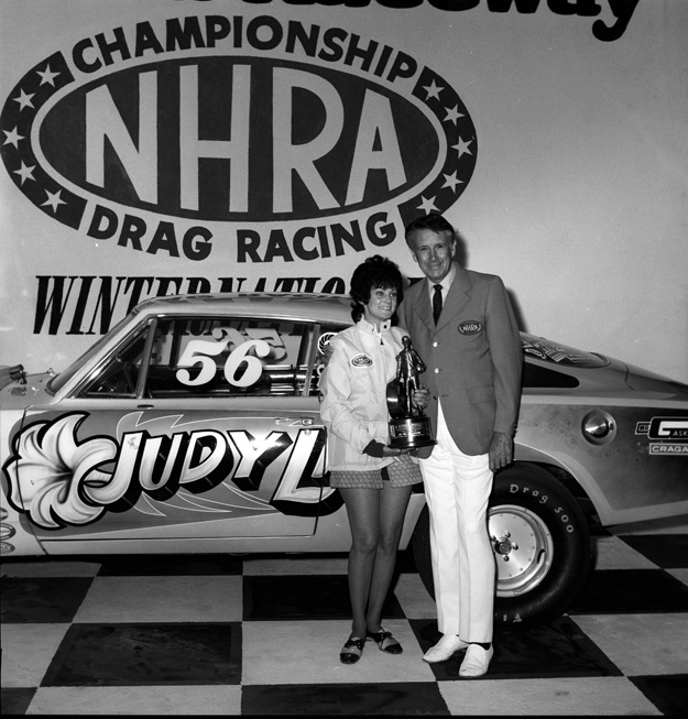 Judy Lilly holding a trophy in front of her drag car