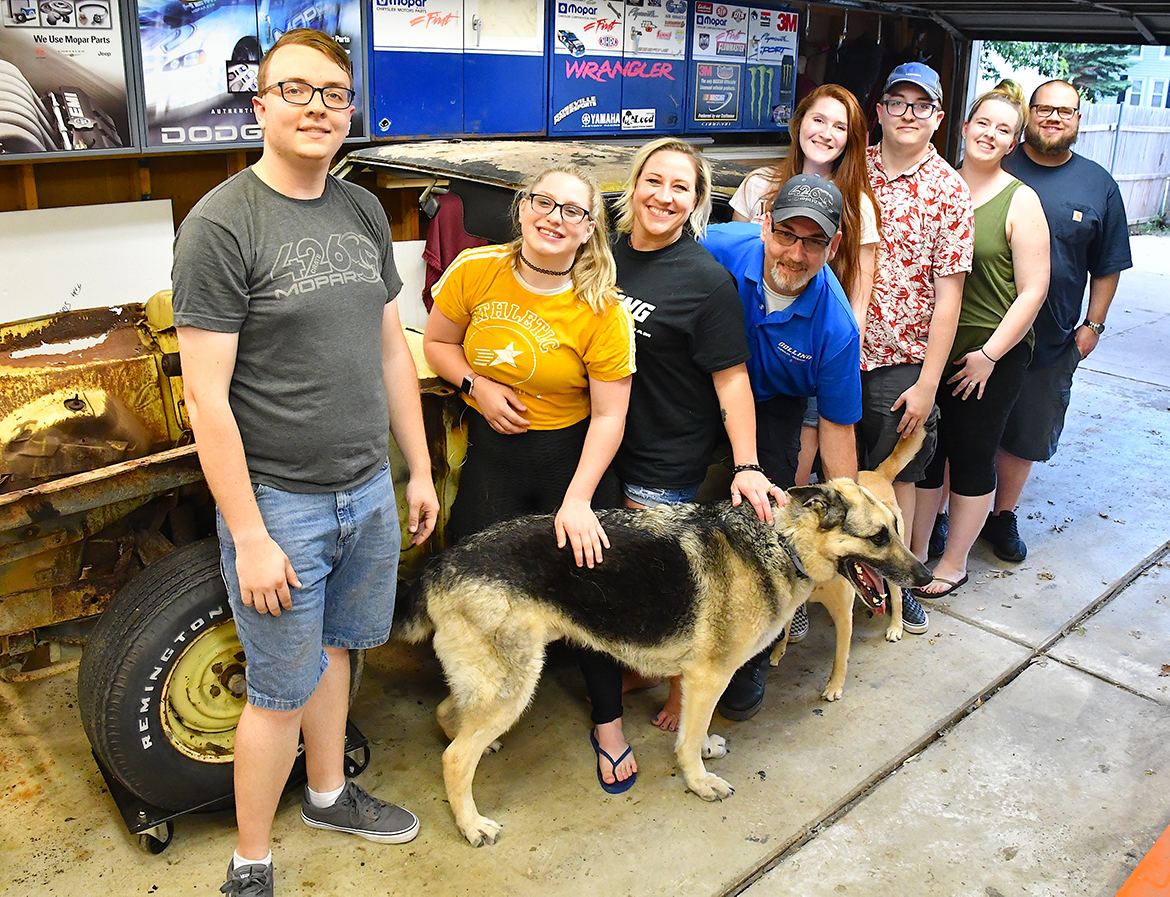 Four men, four women and one dog standing with an old vehicle in a garage