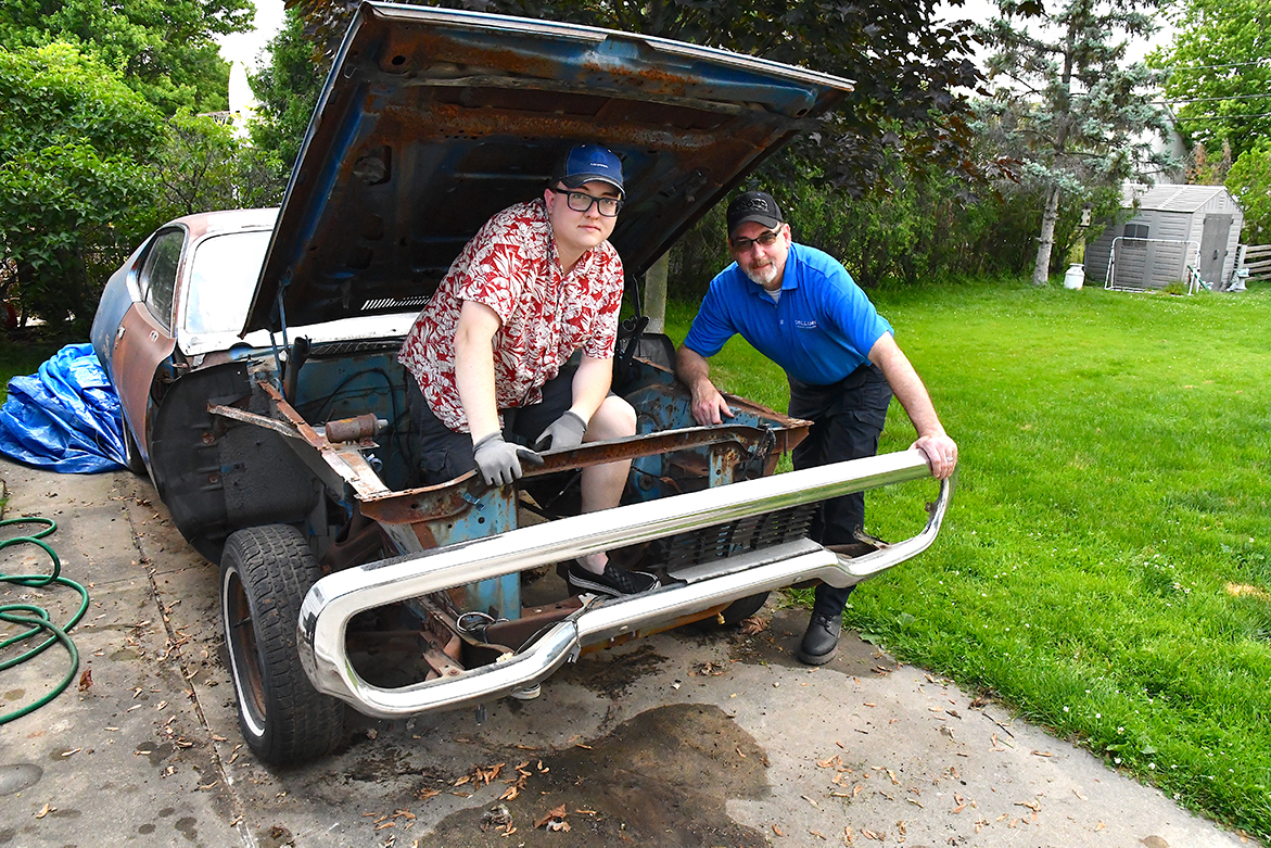 Two men with a rusted vehicle that has parts of the body missing and no engine